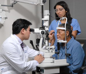 An ophthalmologist examines a person's eye with a lens and slit lamp while another provider holds the patient's head still.