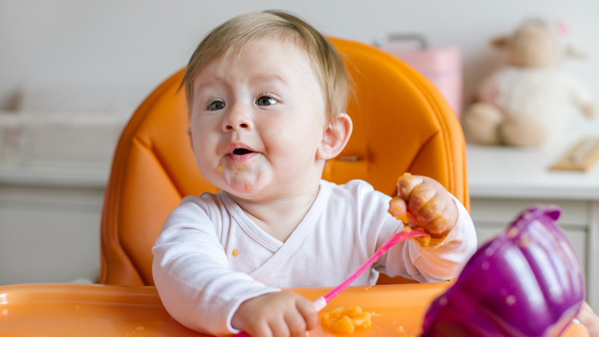 Young baby eats pureed food with hands sitting in high chair.